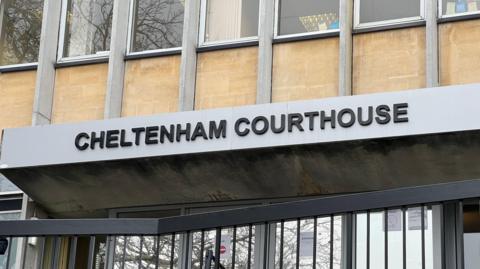 A close-up shot of the entrance at Cheltenham Magistrates' Court. The building is beige with grey panels either side of the windows. "Cheltenham Courthouse" is written above the entrance in capital letters