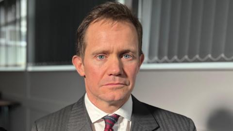 A head and shoulders photo of Mark Teeton, a middle-aged white man with brown hair and piercing light blue eyes, looking into the camera. He wearing a dark grey suit, a white shirt and a red and blue striped tie.
