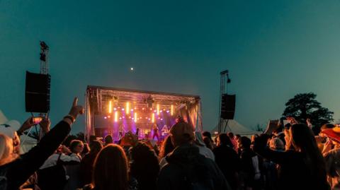 A stage is set up outside and lit up. A crowd has gathered in front of it. The sky is blue.