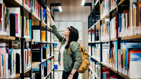 A woman with black hair and a box fringe in a library reaching for a book on a book shelf. She is wearing a green jacket and light brown backpack.