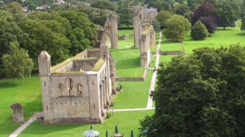 An aerial view of Glastonbury Abbey. It is a large medieval ornate stone structure which has fallen into ruin. Mostly just the frame of the building remains, with several tall spires and arched windows. The grass around it is lush and green and the building is surrounded by mature trees.