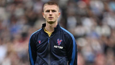 Adam Wharton, wearing a Crystal Palace training top, lining up before a match, with the crowd a blur behind him