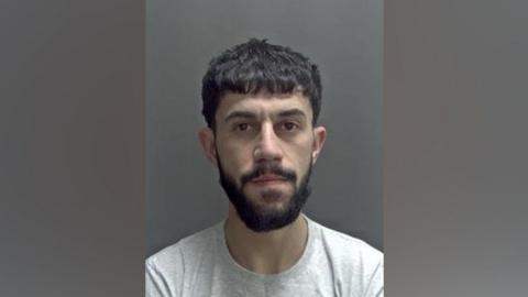 Safa Lawan is looking at the camera in a police custody image. He has dark, short hair and a dark beard and moustache. He is wearing a grey T-shirt