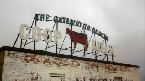 The Co-op cow sign on top of a white painted building. Green lettering says "The Gateway to Health" with white lettering underneath saying "Co-op Milk", and in the middle is a red cow figure.