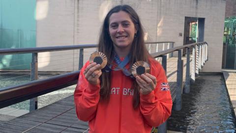 A woman wearing a red Team GB top holds up two bronze Olympic medals with a water feature and other buildings in the background