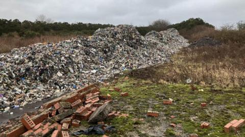 A fly-tipping site, with large piles of household waste, bricks and other rubbish dumped on greenspace
