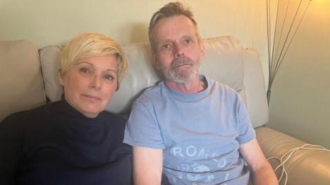 A woman on the left with short blonde hair next to a man with greying hair. They are both sat on a cream leather sofa.
