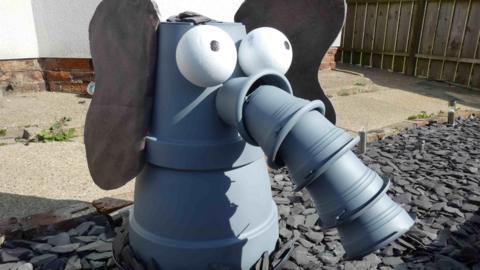 A grey elephant's head made out of plant pots. It is on grey slate in a garden with concrete and a few weeds behind it, a white wall and a wooden fence. 