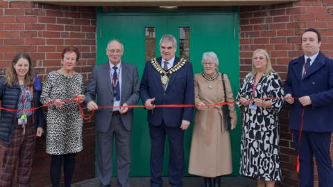 Mayor of Barnsley Cllr John Clarke stands in front of Great Houghton Miners' Welfare Hall, where a red ribbon is being stretched out by attendees for him to cut.