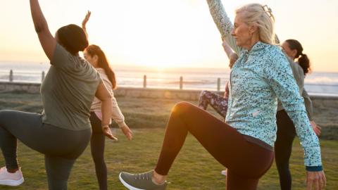 A group of women, standing in a green field at sunrise, wearing exercise clothes and stretching while standing on one foot