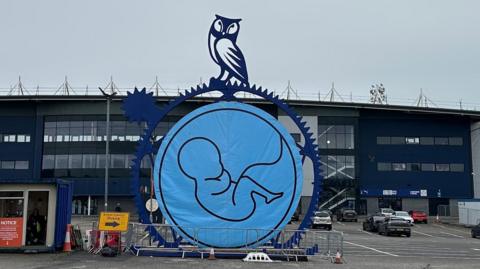 Line drawing of embryonic baby in a blue circle, which is draped inside a sculpture of a cog with the owl mascot of Oldham Athletic above. The work appears outside the car park in front of the stadium.