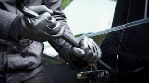 An image of the lower half of a person, dressed in black with black leather gloves, using a crowbar to break into the front door of a black car.