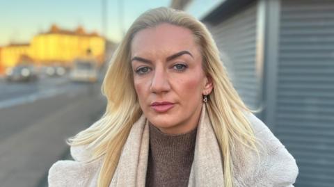 Amy is in her mid-thirties and has long, dyed blonde hair and is wearing a cream coat. She is standing outside next to a shops roller blinds that are out of focus behind her.