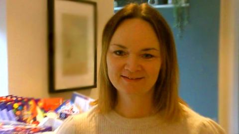 Laura Evans wearing a white jumper and smiling at the camera. She has shoulder-length dark blonde hair and there are presents behind her 