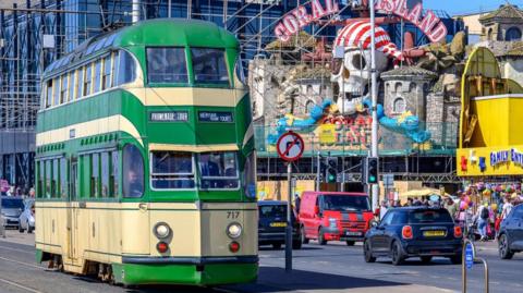 Green and white double-decker Tram 717 which has a unique balloon shape travelling  along Blackpool Promenade during the day with Coral Island in the background. 