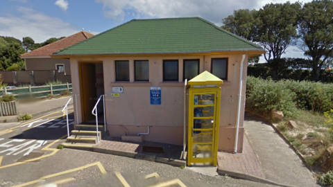 A mid shot of the Green Island public toilets. It is a small hut with a green roof, cream coloured walls, and a yellow phone box outside.