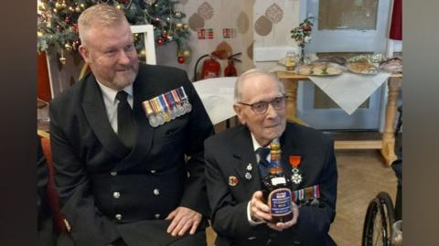 Two men sit next to each other in a room. They are both wearing black jackets with silver buttons, black ties, and white shirts, with several medals pinned to their jackets. The man on the left has greying ginger hair and a beard. The man on the right has grey hair and glasses, and is holding a bottle of rum.