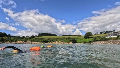 Two swimmers wearing wetsuits and swim hats and towing floats in a river with a grassy area behind them.