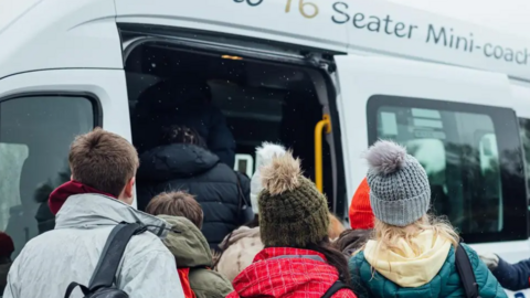 Several boys and girls queue to get on a minibus. They have their backs to the camera and have coats and bobble hats on as some get on at the front of the vehicle.