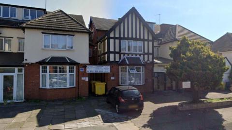 The outside of a red brick house with victorian style crosshatching on the to half. It is a nursing home with a black car parked outside.