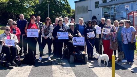 A group of men and women, the majority middle-aged or elderly, stand in the grounds of a leisure centre building in bright sunshine. One woman is sitting on a wheelchair, another in a mobility scooter. One holds a white dog on a lead. Some have placards with slogans reading: "Real trainers, not AI screens", and "Would you take 40% less income."