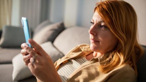 A woman with red hair is sitting on a sofa, holding her smartphone in front of her as she looks directly at its front camera.