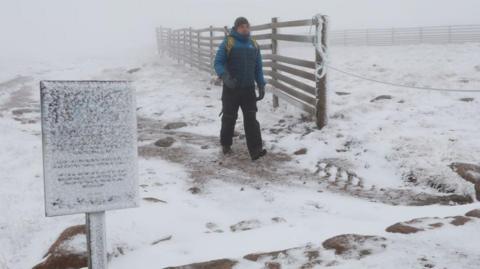 A mountain ranger dressed in winter jacket, hat and boots walking through snow. There is a snow covered sign and wooden fence