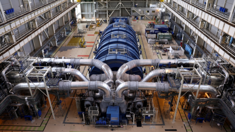 A wide shot of a nuclear reactor. It is a long blue cylinder joined up by many pipes.