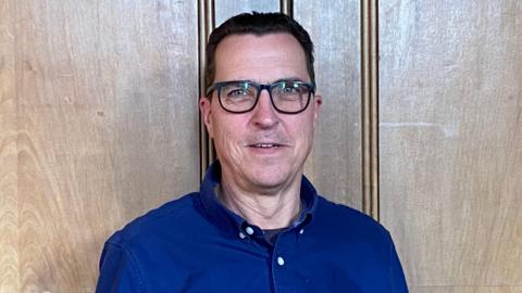 Bob Ensch is standing against a wooden wall. He is smiling at the camera while wearing a blue shirt and blue square framed glasses. He has short black hair and blue eyes. 