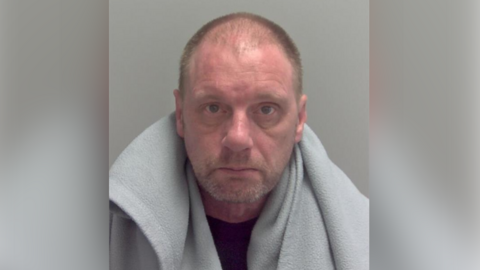 A shaven headed man stares at the camera in a police mugshot. He has a light grey fleecy blanket over his shoulders and is wearing a dark blue T-shirt.