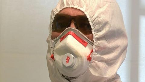 The photo is a close up of trauma cleaner, Jim Gildea's face. There is little skin visible as he's wearing a forensic suit, face mask and protective glasses.