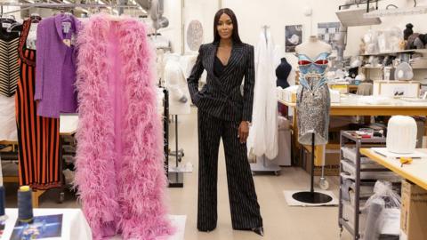 Ms Campbell stands next to her outfits in a design room - with her pink met Gala outfit on the left and a car-inspired metal and plastic corset on the right