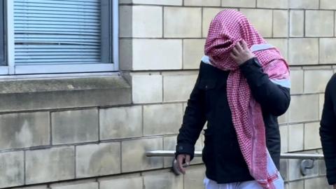 Ashraf Osmani, in a navy jumper with a red and white scarf obscuring his face, walks out of Northampton Crown Court.