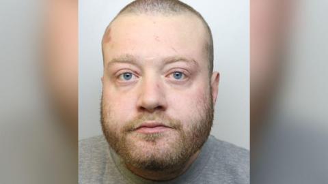 Christopher looks at the camera with a blank expression. He has light hair, shaven very short, and a light beard, and wears a grey prison jumper. 