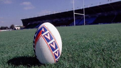 A generic photo of a white rugby ball with red and blue patterning placed upright on green grass, with a stand and white rugby goal beyond.