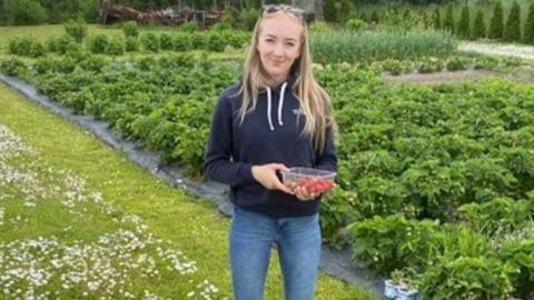 Woman wearing jeans and a navy blue hoodie, she has long blonde hair and sunglasses on top of her head, she is holding a punnet of strawberries and smiling.