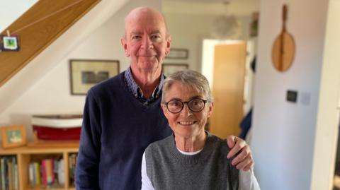Peter is wearing a blue checked shirt with a navy v-neck jumper on top. Helena is wearing a white long sleeve t-shirt with a grey jumper on top. She has black rounded glasses on and has short grey hair. 