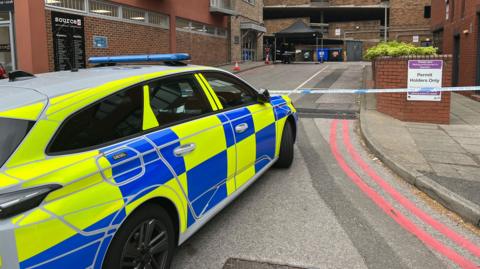 Police tape and traffic cones around the entrance of a building. A police car is parked outside the taped off area.