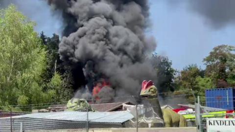 Black smoke and orange flames are seen coming from the unit. There are some model dinosaurs in the foreground behind a barbed wire fence. 