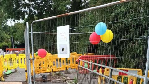 Metal fencing securing an area of the tarmac road and pavement. There is a sinkhole within the fenced off area. There are also green sandbags, yellow plastic barriers and orange traffic cones dotted around to warn people of the danger. There are colourful bunches of balloons tied to the fence.