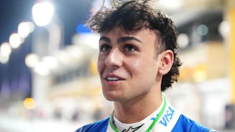 A close up photo of Arvid Lindblad looking up and past the camera. He has long curly brunette hair and is half smiling. He's wearing a blue and white top but you can only see the very top of it in the photo, the background is blurred but it appears like he is on an F1 grid.