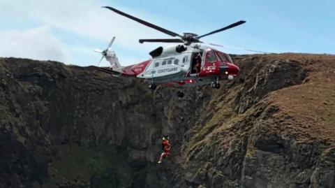 The coastguard helicopter is winching two people very close to a rocky cliff face.
