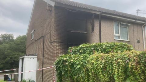 An end-of-terrace property has fire damage around a first floor window. Police tape surrounds the building.