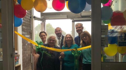 A balloon arch behind automatic doors. There is a yellow ribbon with green bows. Six people are stood behind the ribbon. The woman on the front left is holding scissors ready to cut the ribbon.