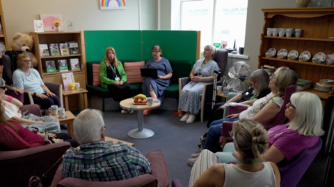 Patients sitting in a circle during one of the group sessions, with a crockery shelf to the right and a round table in the middle with a fruit bowl on it 