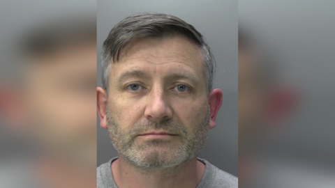A police mugshot of a man with short, cropped grey hair. 