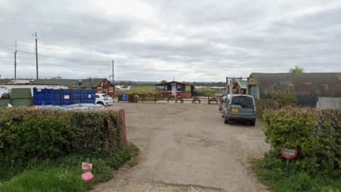 Multiple single storey buildings around a small yard. There are a few cars parked in the yard and some light aircraft can be seen on a grass airfield in the background.