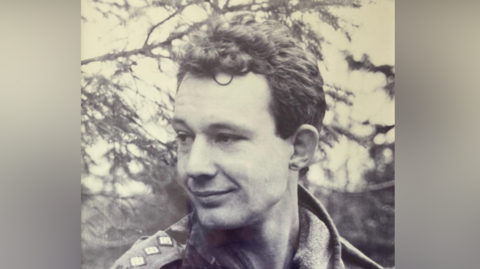 A black and white picture of Major Christopher Dockerty. He is smiling as he looks away from the camera. He has short hair and a shaved beard. He wears an army uniform. 