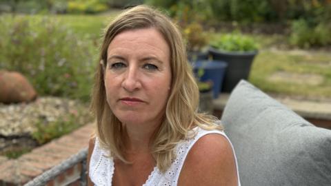 Sadie Hopkins looks towards the camera with a neutral expression. She is wearing a white top and sitting in a garden, there are plant pots and grass visible in the background