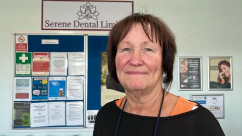 Photo of Mayhew looking into the camera and smiling slightly. She is wearing a black jumper with an orange vest underneath. She has chin-length dark hair, and is stood in the practice waiting room. Behind her, a large sign reads 'Serene Dental', and a blue notice board is covered in brightly coloured posters. 